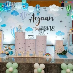 "Blue and white sky-themed first birthday decoration setup by Vruddhi Events, featuring balloon arch, hot air balloons, LED number 1, marquee letters spelling Ayaan, cloud backdrop, and pastel props."