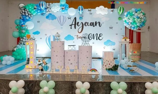 "Blue and white sky-themed first birthday decoration setup by Vruddhi Events, featuring balloon arch, hot air balloons, LED number 1, marquee letters spelling Ayaan, cloud backdrop, and pastel props."