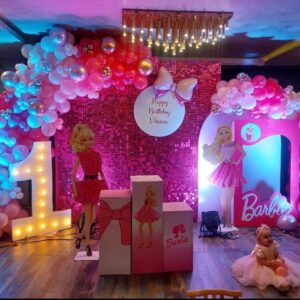 Barbie theme first birthday party decoration with pink balloon arch, LED number 1 light, Barbie doll cutouts, sequin backdrop, and baby girl in a pink dress sitting on the floor – setup by Vruddhi Events in Secunderabad.