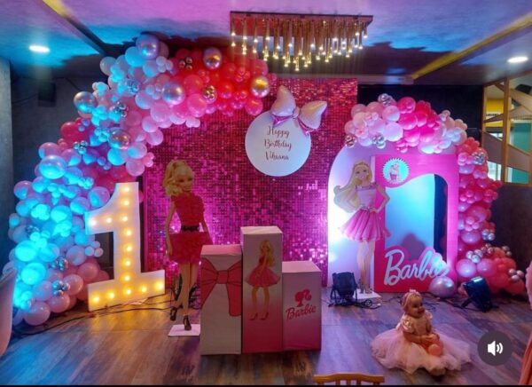 Barbie theme first birthday party decoration with pink balloon arch, LED number 1 light, Barbie doll cutouts, sequin backdrop, and baby girl in a pink dress sitting on the floor – setup by Vruddhi Events in Secunderabad.