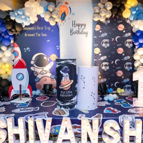Space theme birthday decoration with astronaut and rocket backdrop, balloon arch in black, silver, and blue, constellation props, and illuminated name 'Shivansh' – Vruddhi Events"