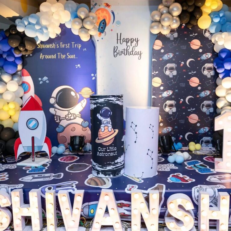 Space theme birthday decoration with astronaut and rocket backdrop, balloon arch in black, silver, and blue, constellation props, and illuminated name 'Shivansh' – Vruddhi Events"