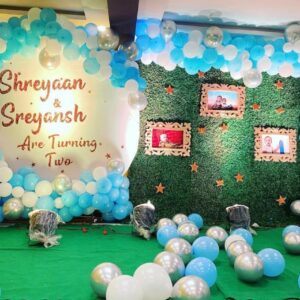 Twin boys' 2nd birthday decoration by Vruddhi Events featuring blue and white balloon arch, green grass wall with framed photos, and silver balloons on the floor for Shreyaan and Sreyansh