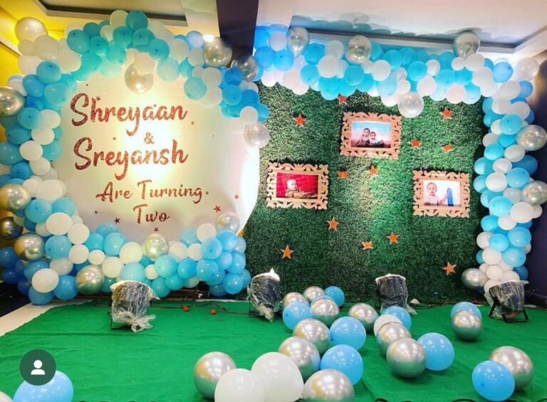 Twin boys' 2nd birthday decoration by Vruddhi Events featuring blue and white balloon arch, green grass wall with framed photos, and silver balloons on the floor for Shreyaan and Sreyansh