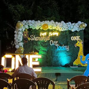 Outdoor jungle safari themed 1st birthday decoration with green backdrop, balloon arch, animal cutouts, light-up 'ONE' letters, and 'Happy Birthday Sharanya' signage
