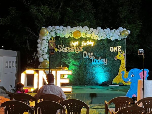 Outdoor jungle safari themed 1st birthday decoration with green backdrop, balloon arch, animal cutouts, light-up 'ONE' letters, and 'Happy Birthday Sharanya' signage