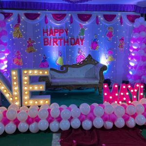 Indoor princess-themed 1st birthday decoration with white and pink balloon arch, cartoon princess cutouts, 'ONE' and 'MAYIKA' light-up letters, and dreamy purple-pink lighting."