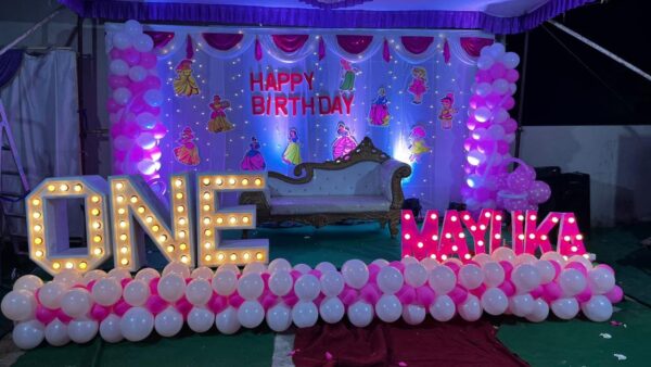 Indoor princess-themed 1st birthday decoration with white and pink balloon arch, cartoon princess cutouts, 'ONE' and 'MAYIKA' light-up letters, and dreamy purple-pink lighting."
