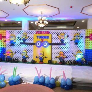 Minions-themed birthday stage decoration with colorful balloon backdrop, 3D Minion figures, and custom name board reading 'Vihaan Turns 2' in a banquet hall setting.