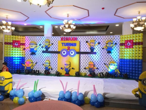 Minions-themed birthday stage decoration with colorful balloon backdrop, 3D Minion figures, and custom name board reading 'Vihaan Turns 2' in a banquet hall setting.