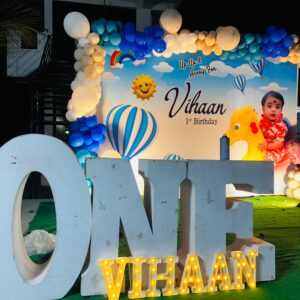 Hot Air Balloon themed 1st birthday decoration for baby boy named Vihaan, featuring a colorful balloon arch, custom backdrop with clouds, sun, and hot air balloons, large 'ONE' letters with Vihaan's name in LED lights, and a cute baby photo on the backdrop