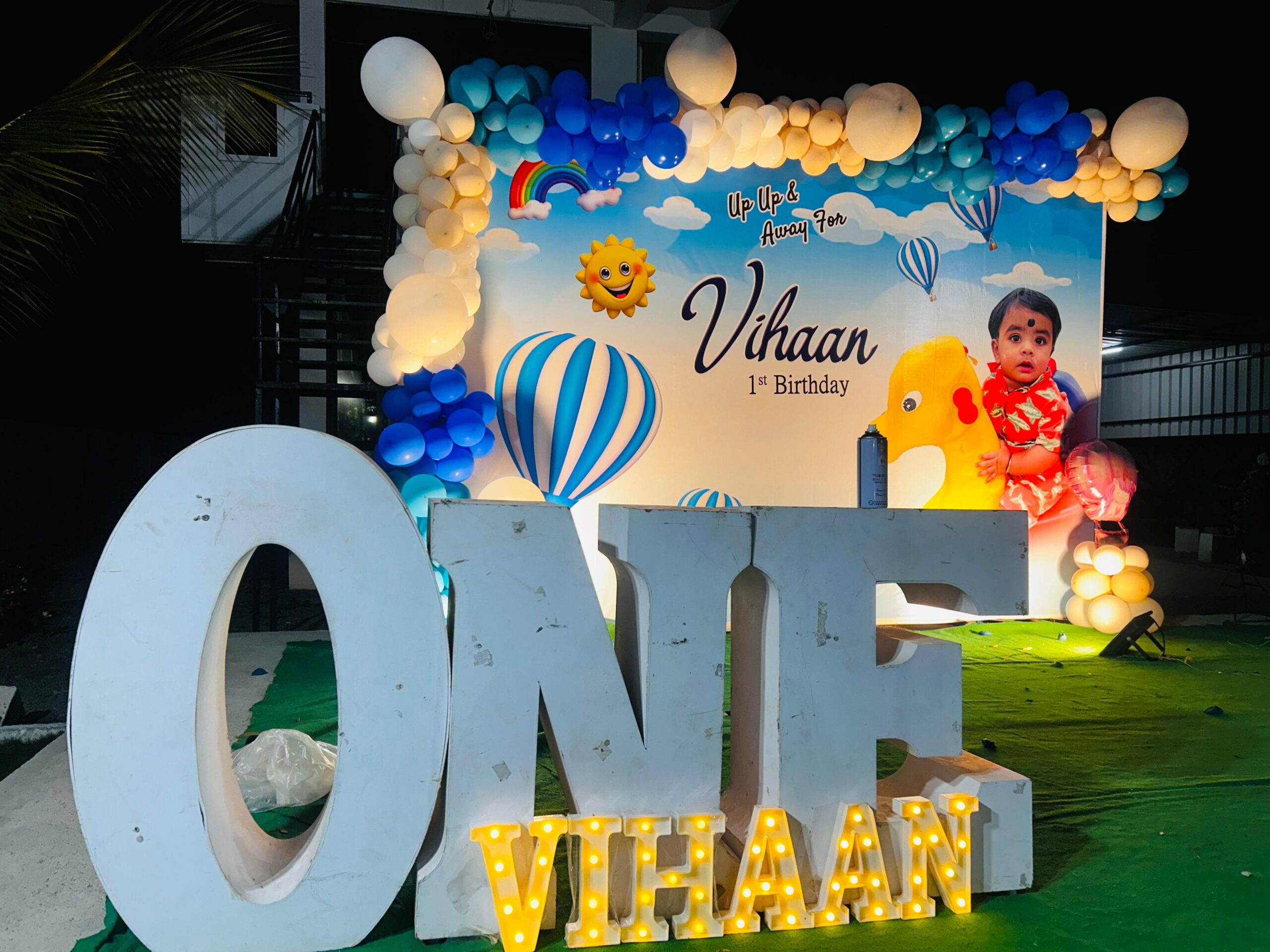 Hot Air Balloon themed 1st birthday decoration for baby boy named Vihaan, featuring a colorful balloon arch, custom backdrop with clouds, sun, and hot air balloons, large 'ONE' letters with Vihaan's name in LED lights, and a cute baby photo on the backdrop