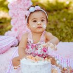 A charming baby girl delights in a cake smash during an outdoor picnic birthday celebration surrounded by pink decor and greenery.