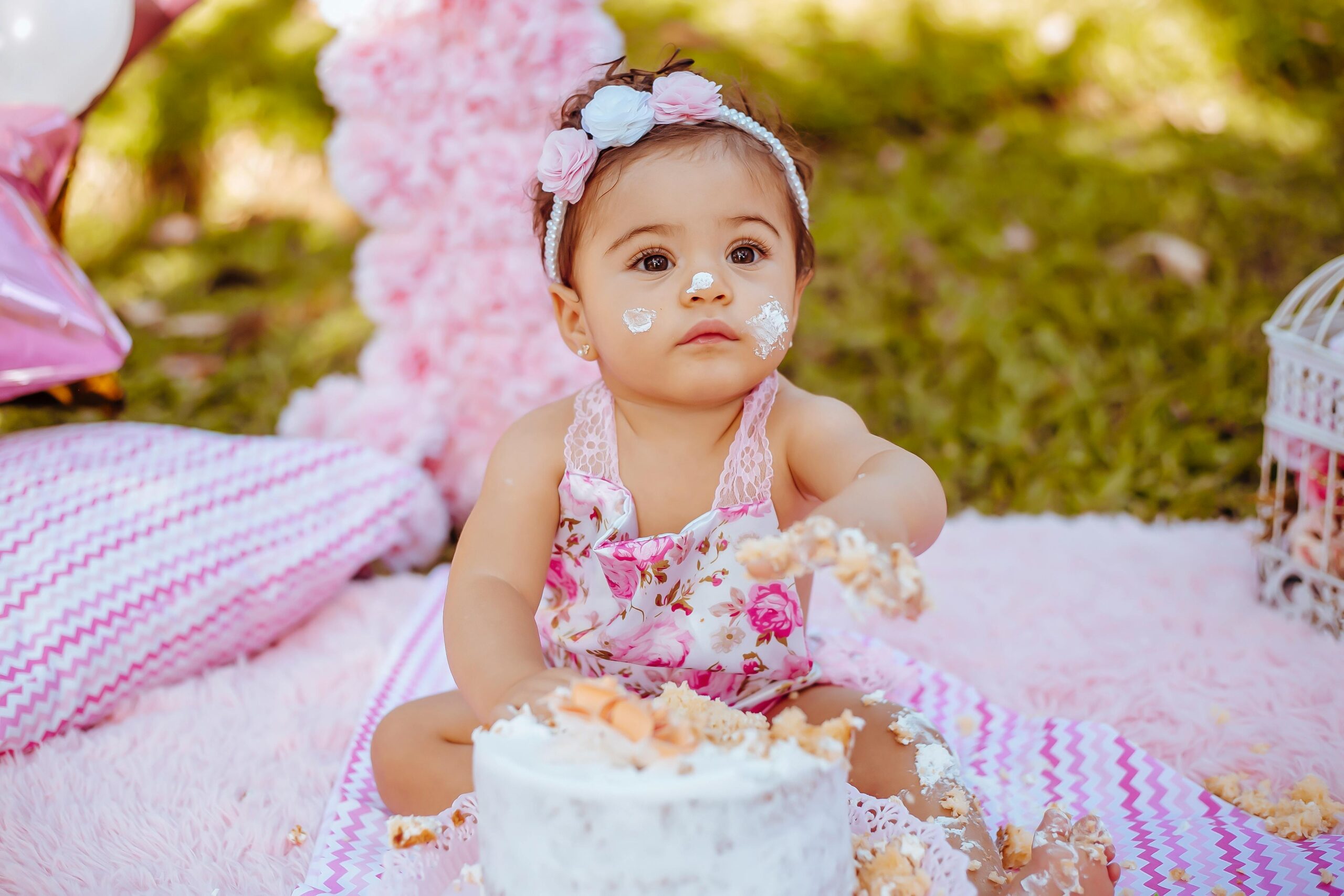 A charming baby girl delights in a cake smash during an outdoor picnic birthday celebration surrounded by pink decor and greenery.