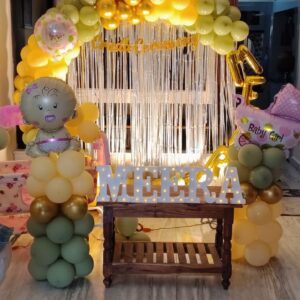 Luxury baby girl welcome balloon decoration with pastel yellow and sage green organic balloon arch, silver shimmer backdrop, LED name letters, baby girl foil balloons, and premium home welcome setup by Pushpaa Party Hub in Hyderabad."